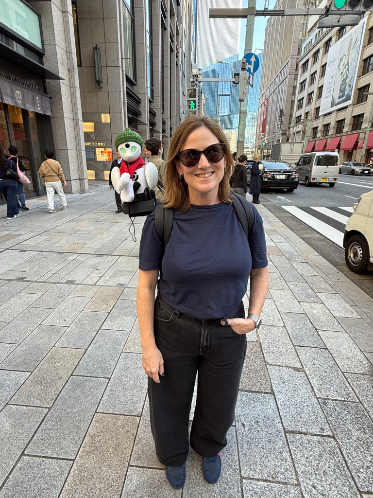 A woman stands on a city street with a palm-sized robot on her shoulder, set against a backdrop of modern buildings.