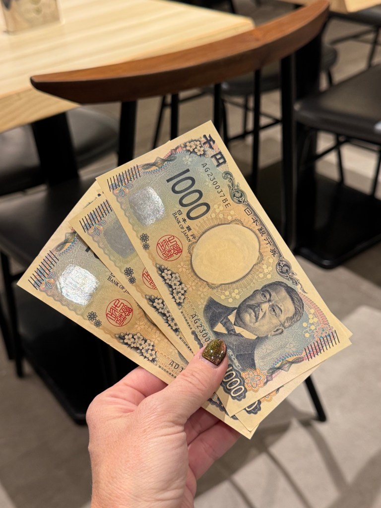 A hand holding three 1000 yen banknotes against a backdrop of a wooden table and chairs.