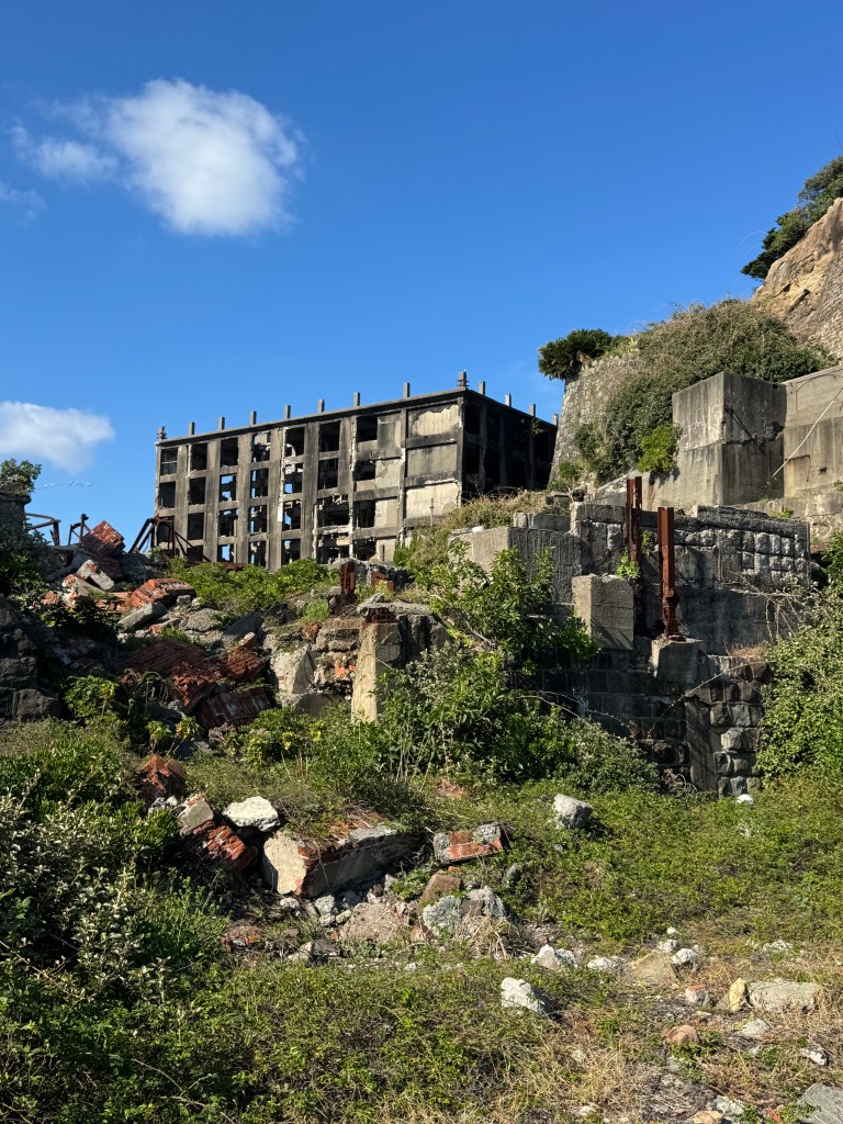 A view of the crumbling remains of a building on Gunkanjima, with overgrown vegetation and partly collapsed concrete structures under a blue sky.
