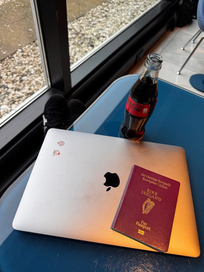 A laptop with stickers on it is placed on a table next to a bottle of Coca-Cola and an Irish passport, with a view of a pebble-strewn ground outside.
