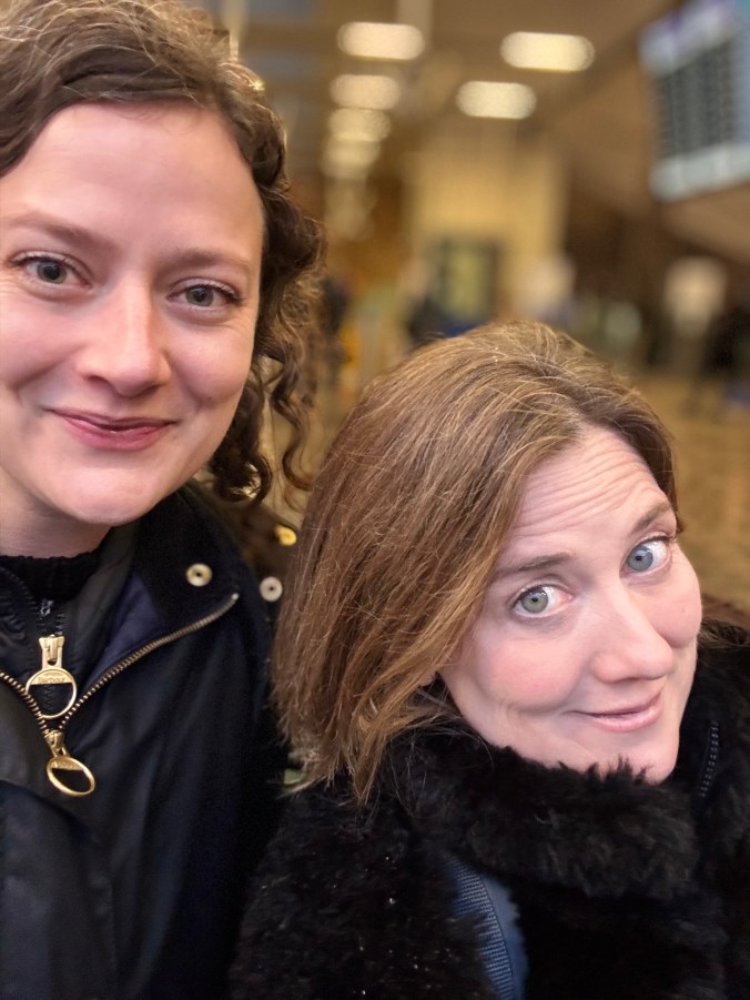 A close-up selfie of two women smiling at the camera, with a blurred background of bright overhead lights.