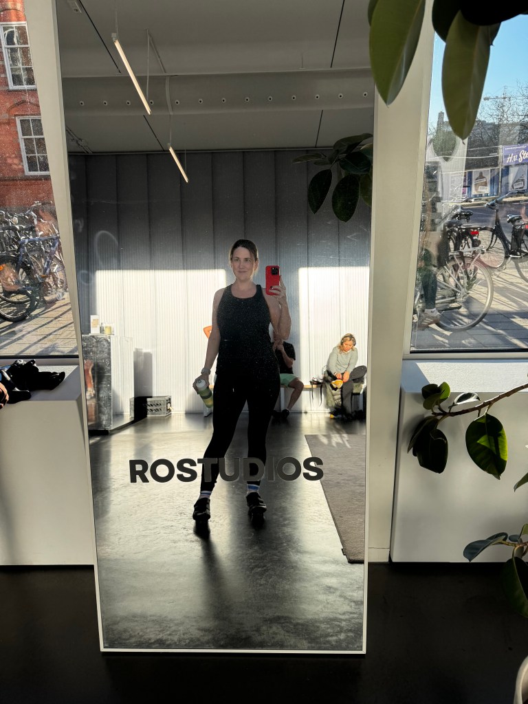 A mirror reflection of a person in athletic wear holding a water bottle, standing in a fitness studio with a sign that reads 'ROSTUDIOS.' Various fitness equipment and other individuals are visible in the background.