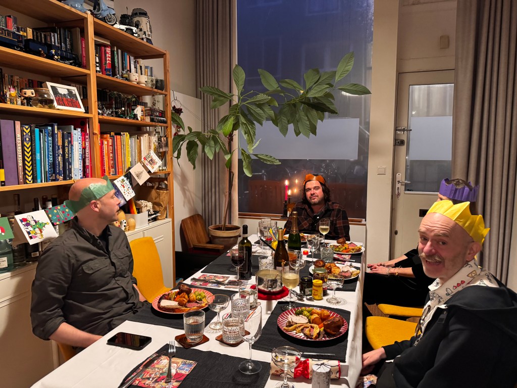 A festive dining table set for a holiday meal with three men wearing paper crowns, surrounded by food and drinks, and a bookshelf in the background.