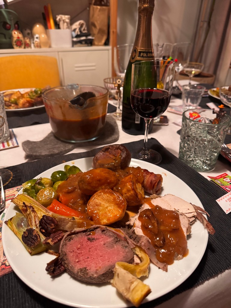 A festive dinner plate featuring various cooked meats, roasted potatoes, Brussels sprouts, carrots, and gravy, with glasses of red wine and champagne in the background.