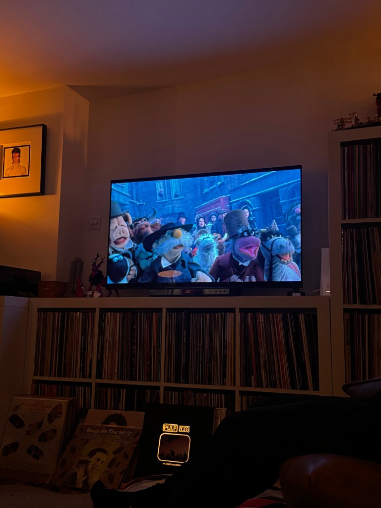 A cozy living room scene featuring a television displaying a scene from 'The Muppets Christmas Carol', with a collection of vinyl records visible in the background.