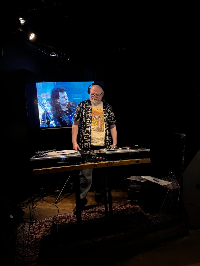 A DJ stands in front of turntables, wearing a patterned shirt and glasses, with a large screen displaying a musician in the background.
