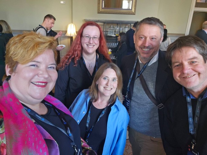A group of five professionals posing for a photo at an event, smiling and wearing lanyards, with a background featuring a stylish interior.