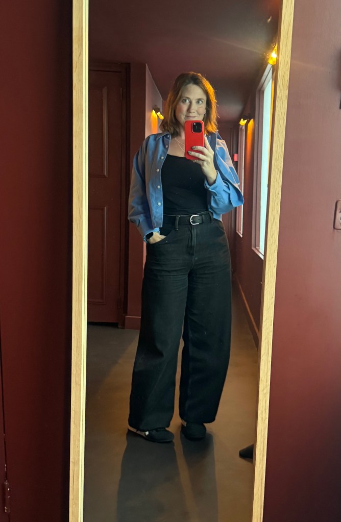 Sharon O'Dea standing in front of a mirror, wearing a blue button-up shirt over a black top and black wide-leg pants, holding a phone for a selfie in a dimly lit hallway.