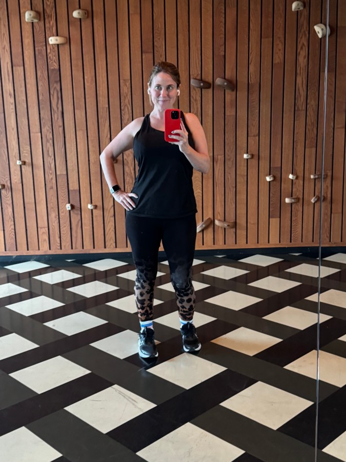 A person standing in a fitness studio, wearing athletic clothing and taking a selfie in the mirror.