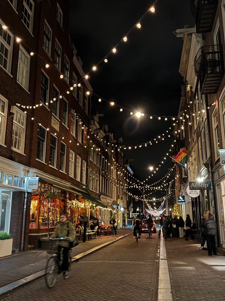 A picturesque street scene at night, illuminated by string lights and decorated storefronts. A cyclist is riding down the cobbled street, with people dining outside and an inviting atmosphere created by warm lighting.