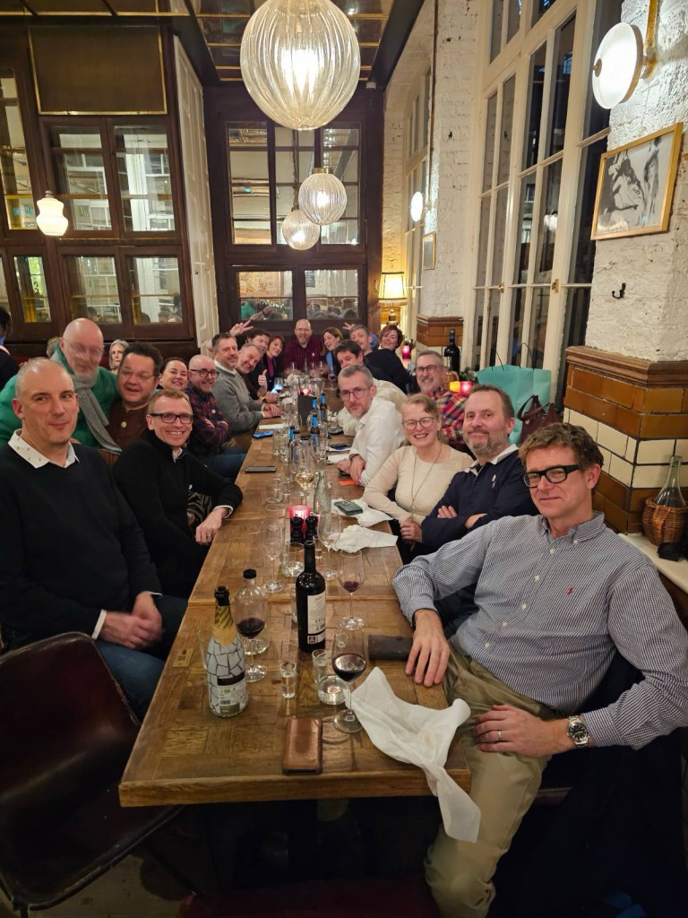 A large group of people gathered around a long wooden table at a restaurant, smiling and enjoying a festive dinner together, with glasses and plates scattered on the table.
