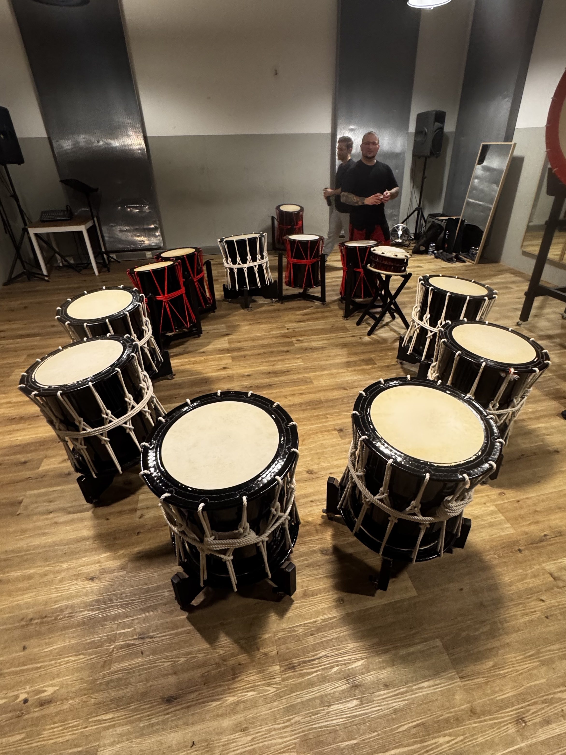 A group of traditional drums arranged in a circle on a wooden floor, with two individuals visible in the background. The setting appears to be a rehearsal space.