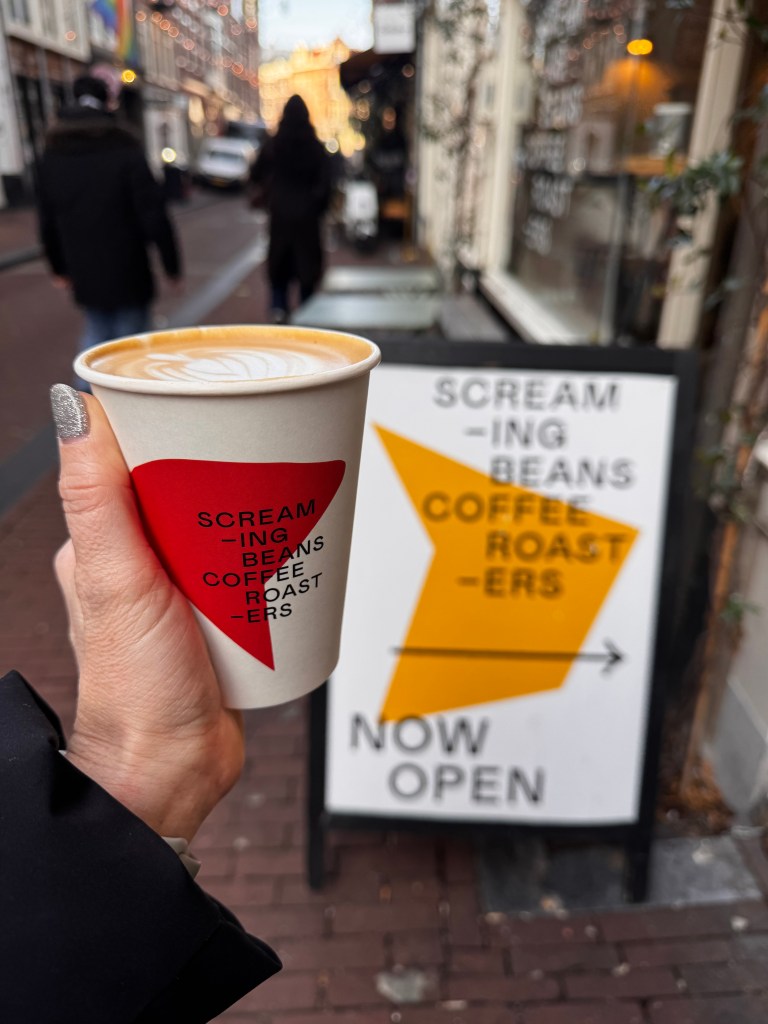 Hand holding a coffee cup with a red and white design in front of a coffee shop sign that reads 'SCREAM-ING BEANS COFFEE ROAST-ERS NOW OPEN'.