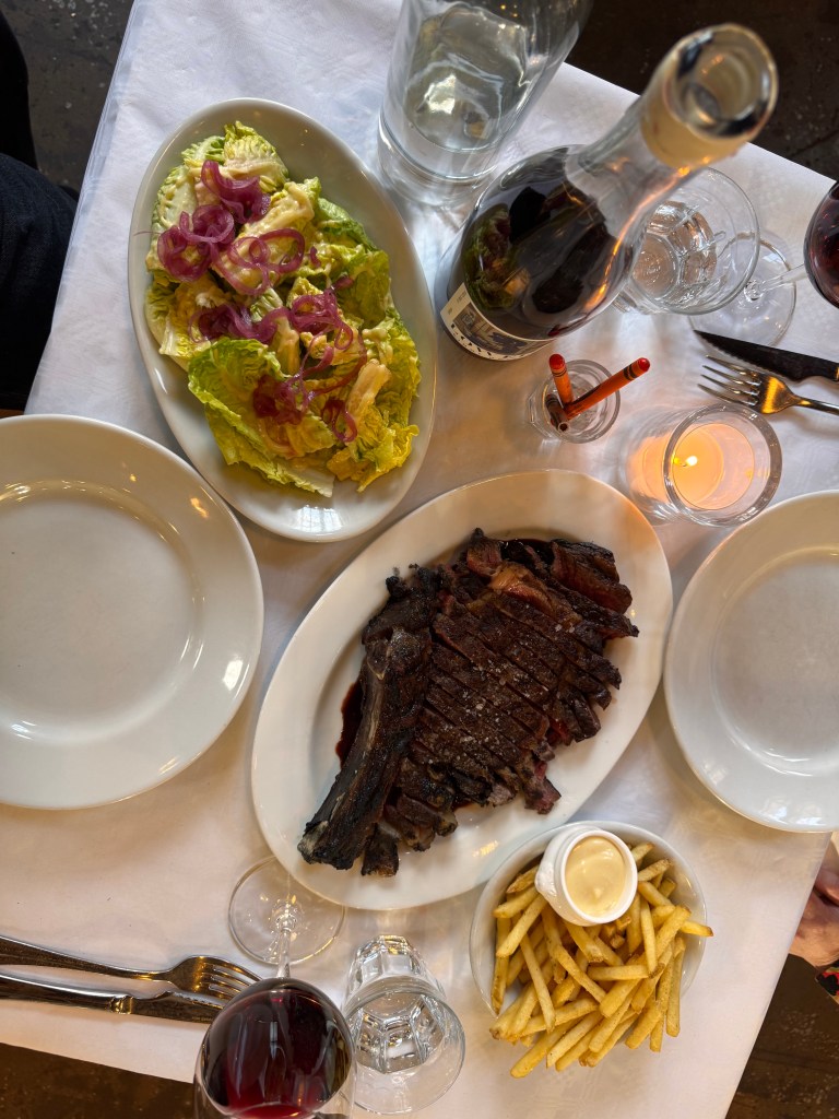 Table setting featuring a plate of grilled steak, a side salad with lettuce and pickled onions, crispy French fries with dip, a bottle of red wine, wine glasses, and a glass of water.