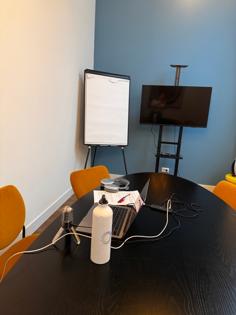 A meeting room setup featuring a laptop, microphone, water bottle, and stationery on a dark wooden table, with an empty flip chart and a television in the background.