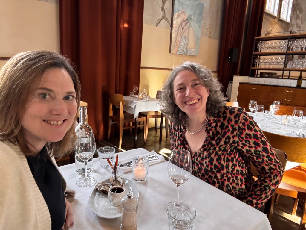 Two women smiling and seated at a restaurant table with a white tablecloth, featuring glassware and a small dish of food in front of them.