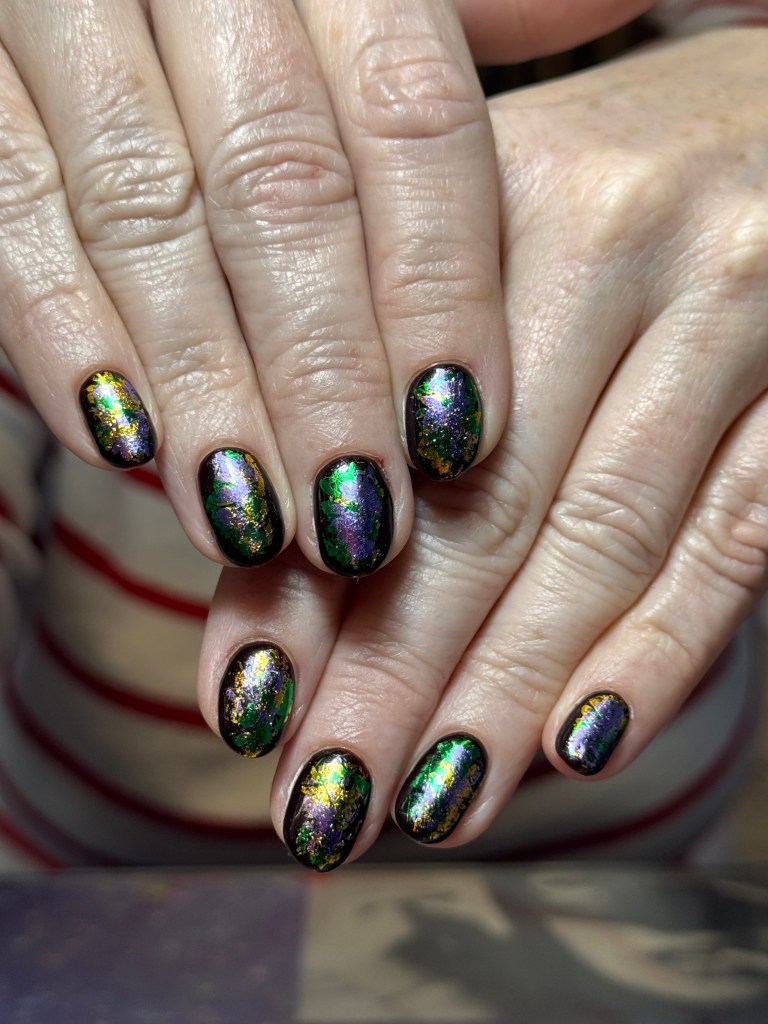 Close-up of a person's hands showing manicured nails with a multicolored, iridescent finish featuring shades of purple, green, and gold.