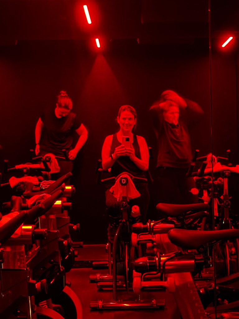 People in a gym studio with red lighting, preparing for a cycling class. One person is taking a selfie while two others are adjusting their equipment.