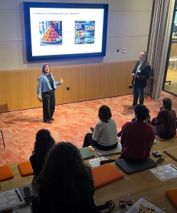 A woman presents in front of a screen displaying images of food, discussing the concept of 'infobesity', while an audience sits attentively in a meeting space.