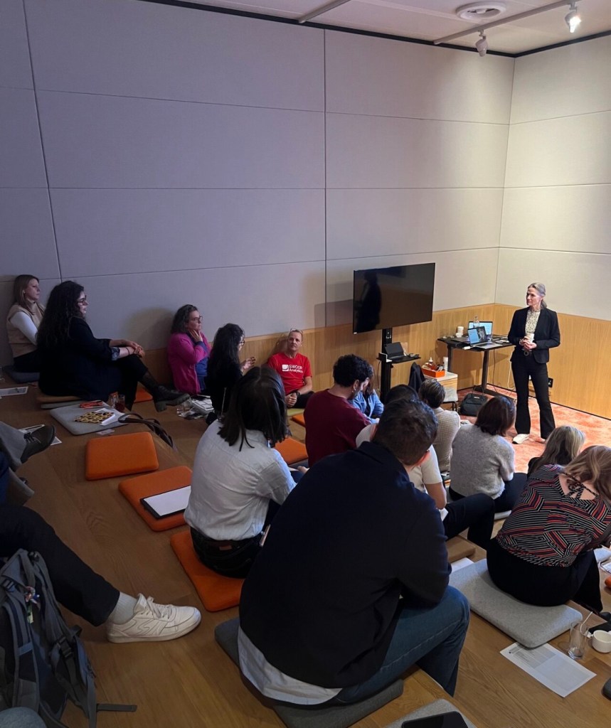 A presenter stands in front of an audience seated on steps in a modern seminar space, with a TV screen displaying a presentation.