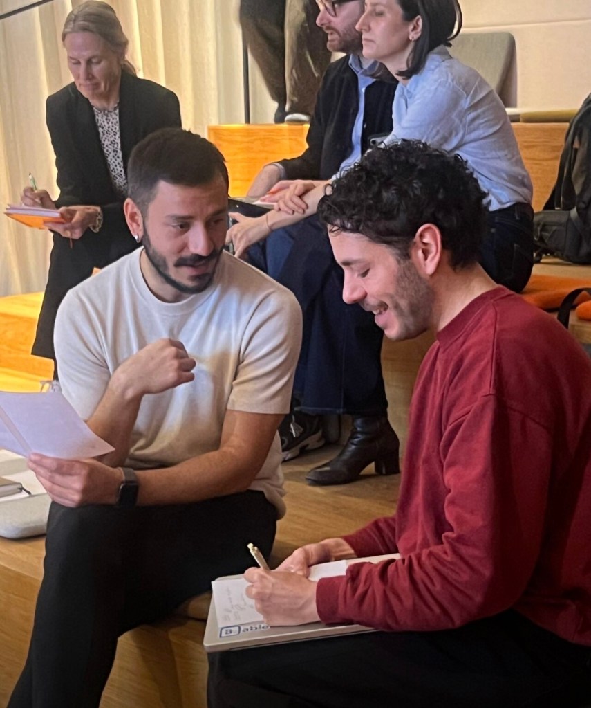 Two men engaged in a discussion, one holding a sheet of paper and the other writing in a notebook, while others are seated in the background.