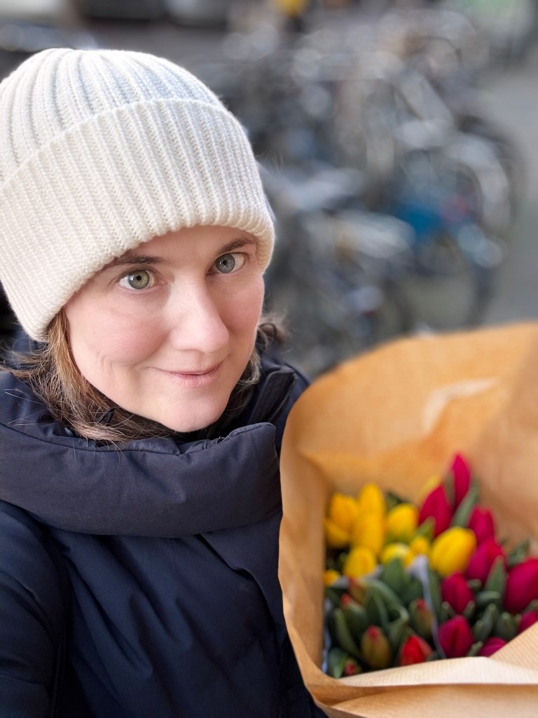 A woman wearing a cream beanie and a black jacket holds a bouquet of colorful tulips, including red and yellow flowers, while standing in front of bicycles.