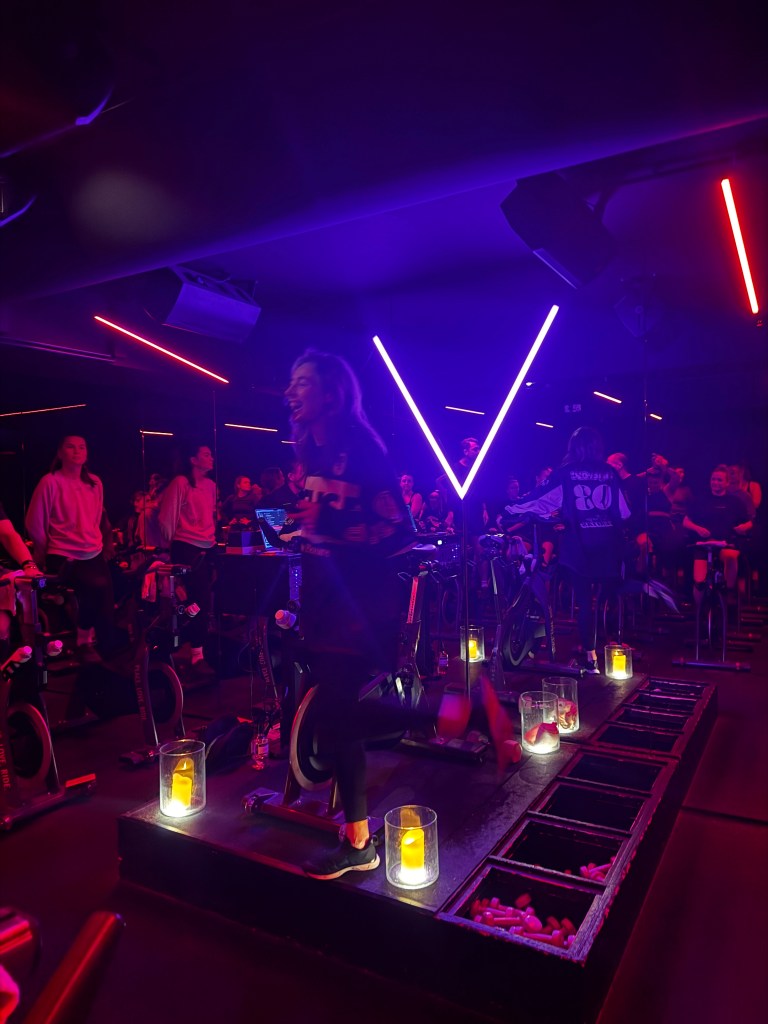 Indoor cycling class with dim lighting, featuring a vibrant purple and red neon 'V' light, and participants on stationary bikes surrounded by glowing candles.