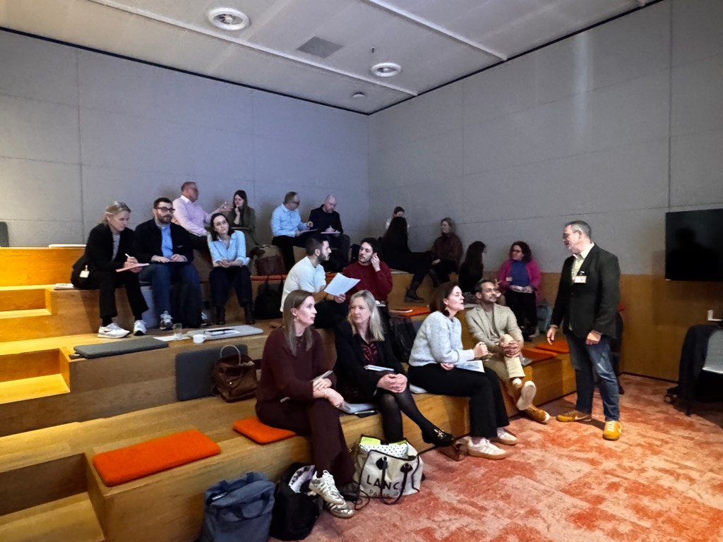A group of people seated in a modern meeting space with tiered seating, engaged in discussions and note-taking.