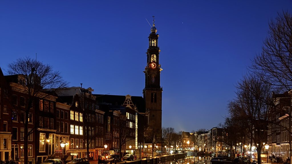 Night view of a historic clock tower beside a canal, with illuminated buildings and street lamps in Amsterdam.
