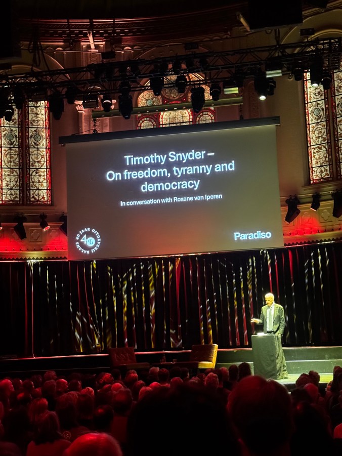 A speaker, Timothy Snyder, is presenting on the topics of freedom, tyranny, and democracy at an event in Paradiso, with Roxane van Iperen in conversation. The background features a large screen displaying the event details and an audience is visible in the foreground.