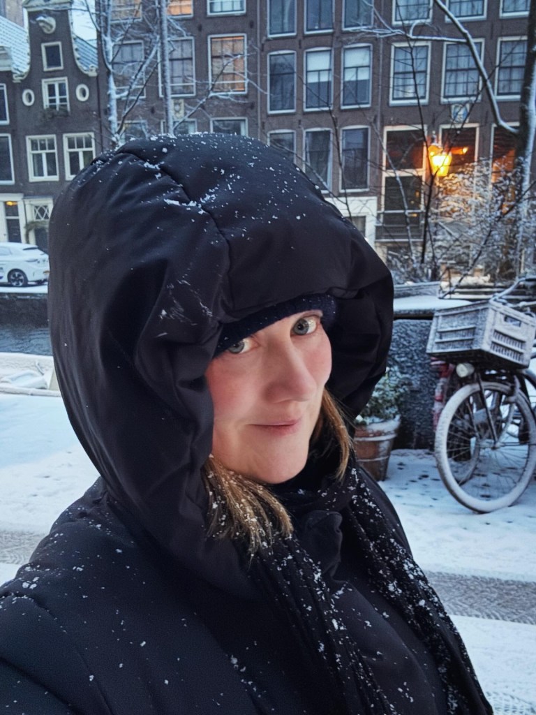 A person wearing a black puffer jacket with a hood, standing in a snowy urban environment with snowflakes visible in the air. Buildings can be seen in the background, along with a bicycle and snow-covered ground.