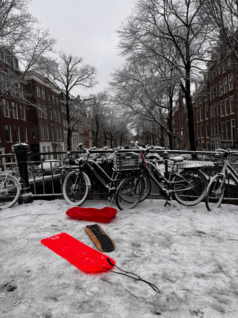 A winter scene featuring a snow-covered street in Amsterdam, with bicycles parked along a canal. Two colorful sleds, one red and one orange, are prominently placed on the snowy ground.