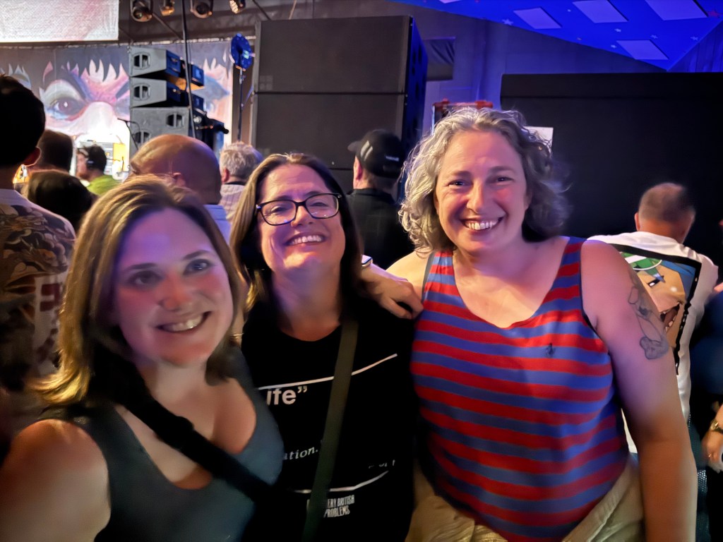 Three women smiling and posing together at a lively event, with a colorful background and concert-like atmosphere.