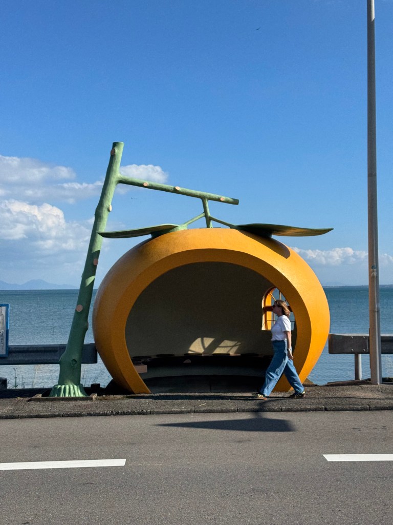 A woman walking past a large, brightly colored bus stop shaped like an orange, with a lake and blue sky in the background.