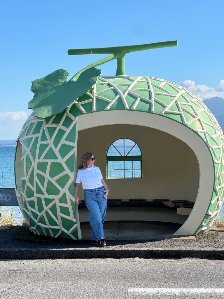 A person leans casually against a large green and white melon-shaped structure that serves as a bus stop, with a scenic waterfront view in the background.