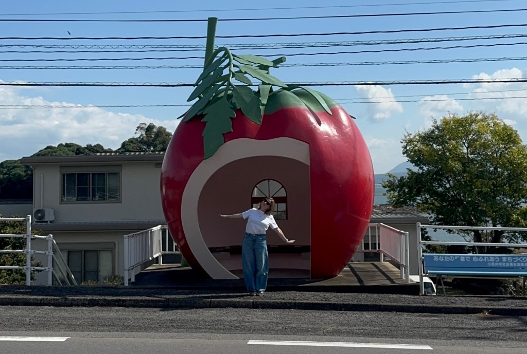 A person standing in front of a large tomato-shaped structure with a smiling expression, wearing a white shirt and blue jeans, against a blue sky with clouds.