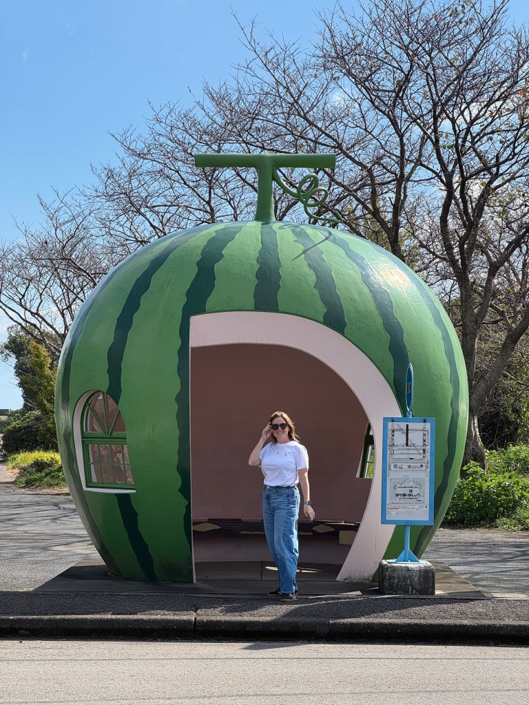 A woman standing next to a whimsical bus stop designed to resemble a watermelon, with bare trees and a blue sky in the background.