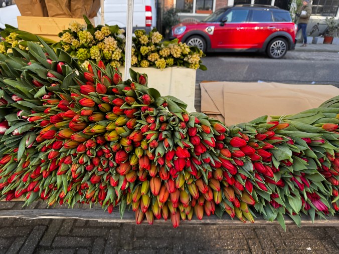 A vibrant display of red and green tulips stacked on a market table, with a blurred background featuring a red car and boxes of flowers.