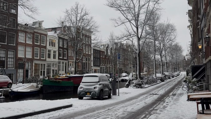 Snow-covered street in Amsterdam featuring traditional Dutch architecture, parked cars, bicycles, and a canal lined with boats.