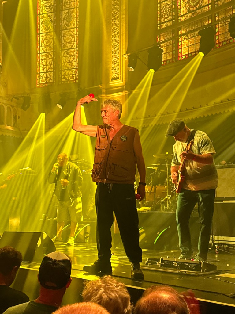 A performer on stage wearing a sleeveless vest and holding a red object, surrounded by yellow stage lights and musicians in the background.