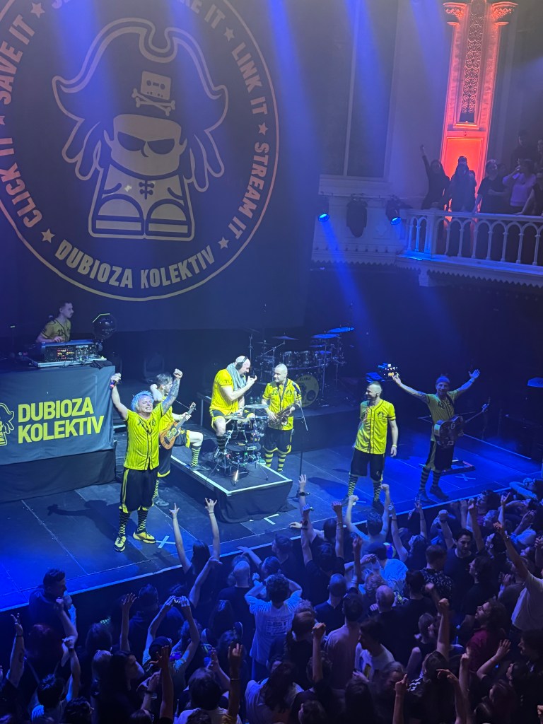 A live performance by the band Dubioza Kolektiv, featuring members in matching yellow outfits, engaging with an enthusiastic crowd in front of them. The stage is illuminated with colorful lights, and a large backdrop of the band's logo is visible.
