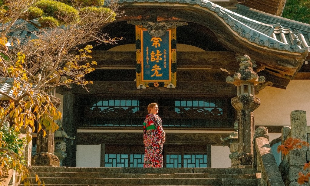 A woman wearing a colorful traditional kimono stands on steps leading to a temple, surrounded by autumn foliage and architecture.