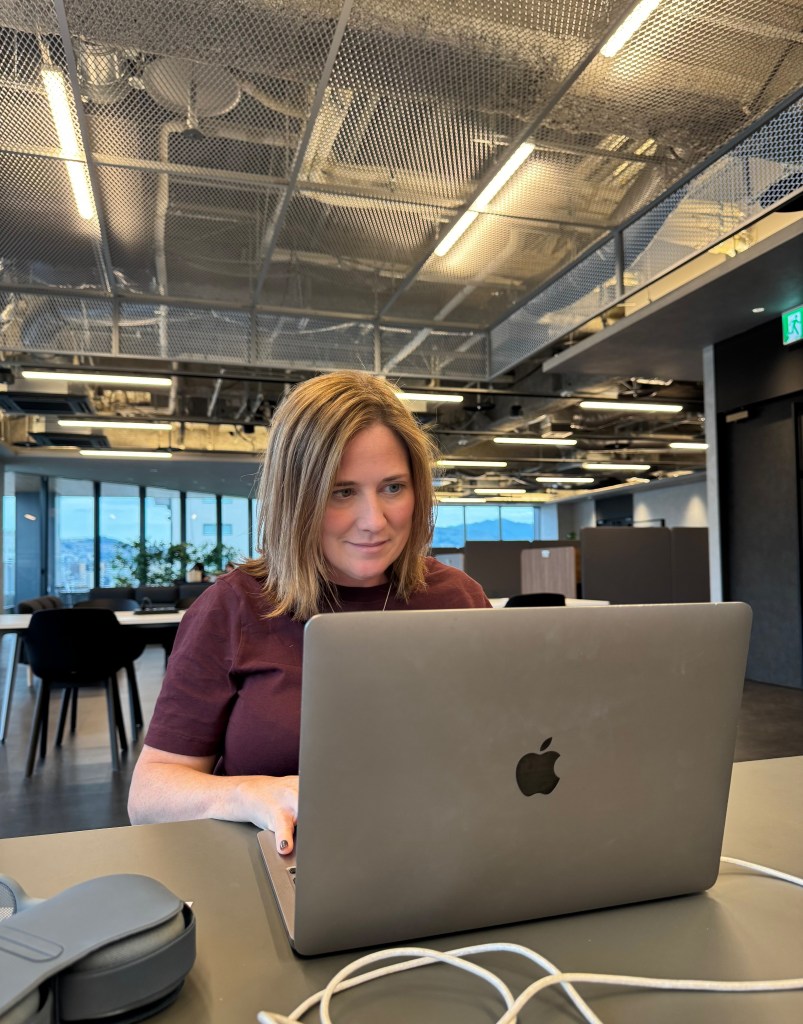 A person with shoulder-length brown hair is sitting at a desk, focused on a laptop in a modern office space with large windows showing a cityscape in the background.