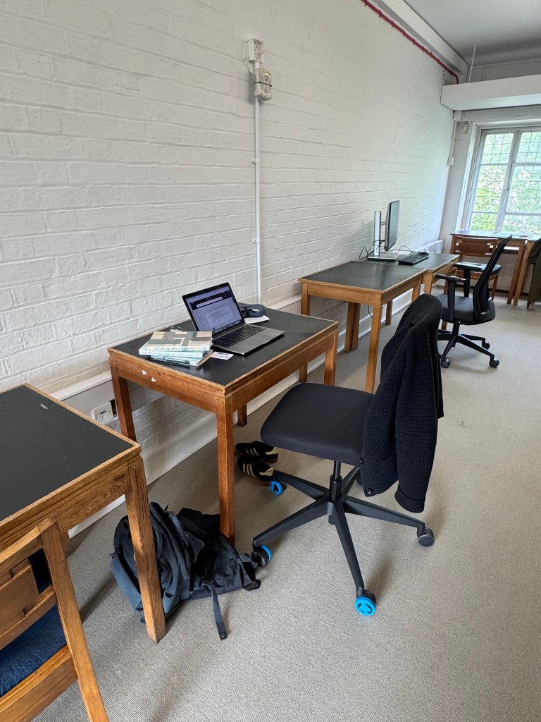 A workspace featuring wooden tables, a laptop, and a desktop computer against a white brick wall, with a bag and a jacket on the floor.