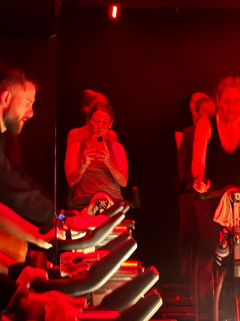 A group of people participating in a spin class, with a focus on one woman taking a selfie in a dimly lit, red-tinted environment.