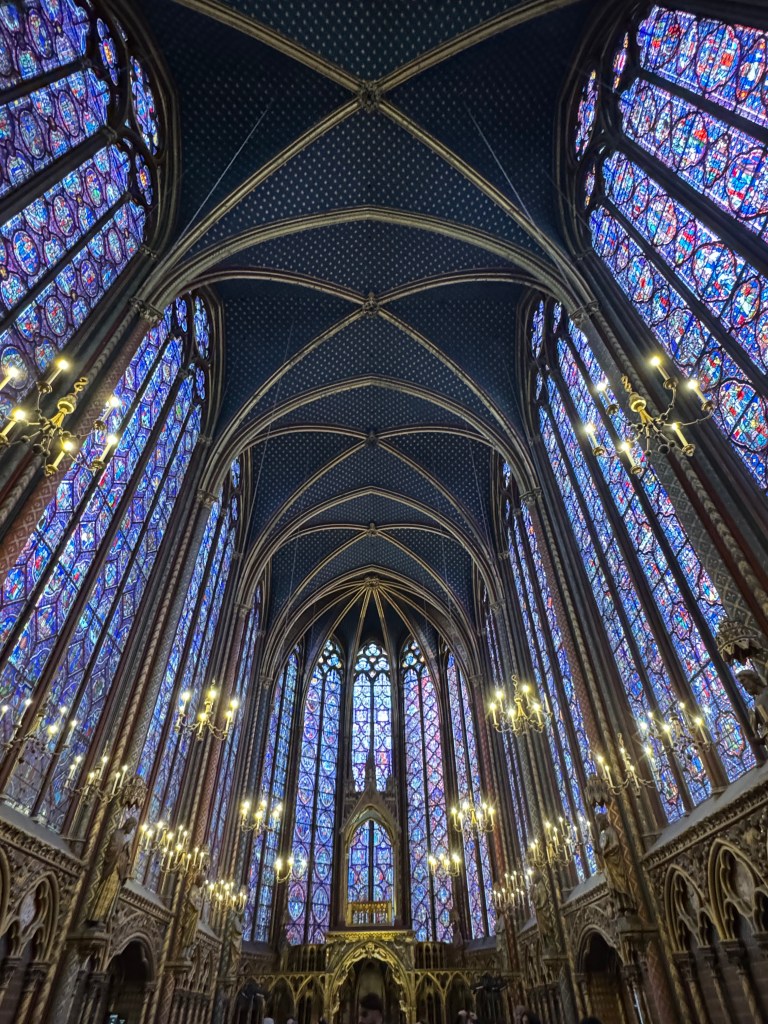 Interior view of the Sainte-Chapelle in Paris, showcasing ornate stained glass windows, a high vaulted ceiling, and chandeliers, creating a colorful and serene atmosphere.