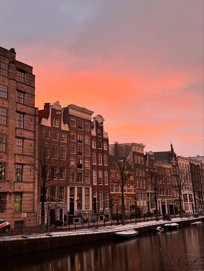 Sunset over a canal in Amsterdam, featuring historic buildings along the water's edge, with a pink and orange sky reflecting on the calm water.