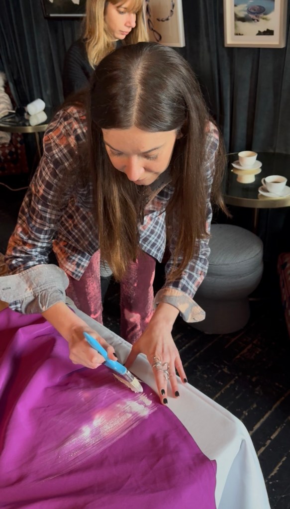 A woman closely examines a purple fabric while applying something with a brush, focused on her work, in a stylish indoor setting. Another person is visible in the background.