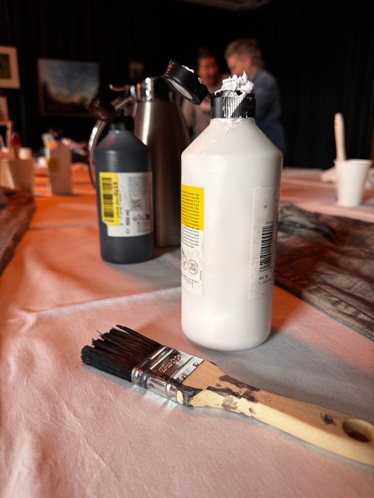 A paintbrush rests on a table next to a bottle of white paint and a spray bottle, with a blurred background of people engaging in an art activity.