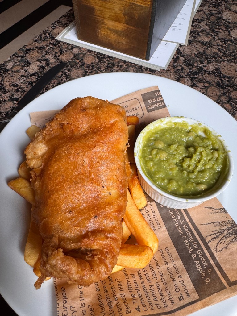 A plate of fish and chips featuring a large, golden-battered piece of fried fish served over crispy French fries, accompanied by a small bowl of mushy peas.